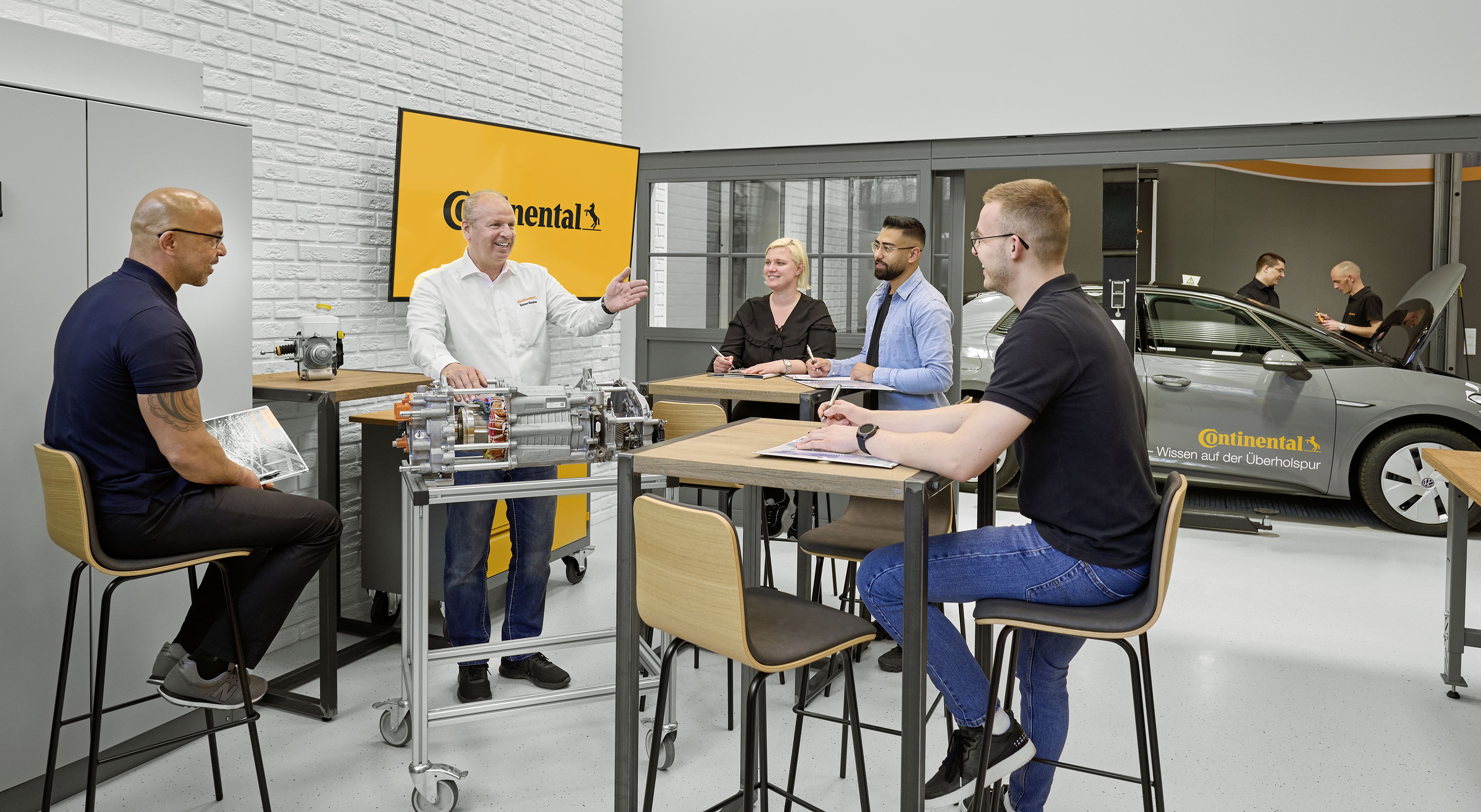 Mitarbeiter hält mit einer vierköpfigen gemischten Gruppe ein Training ab. Im Hintergrund befindet sich ein graues Fahrzeug mit dem Continental Schriftzug.