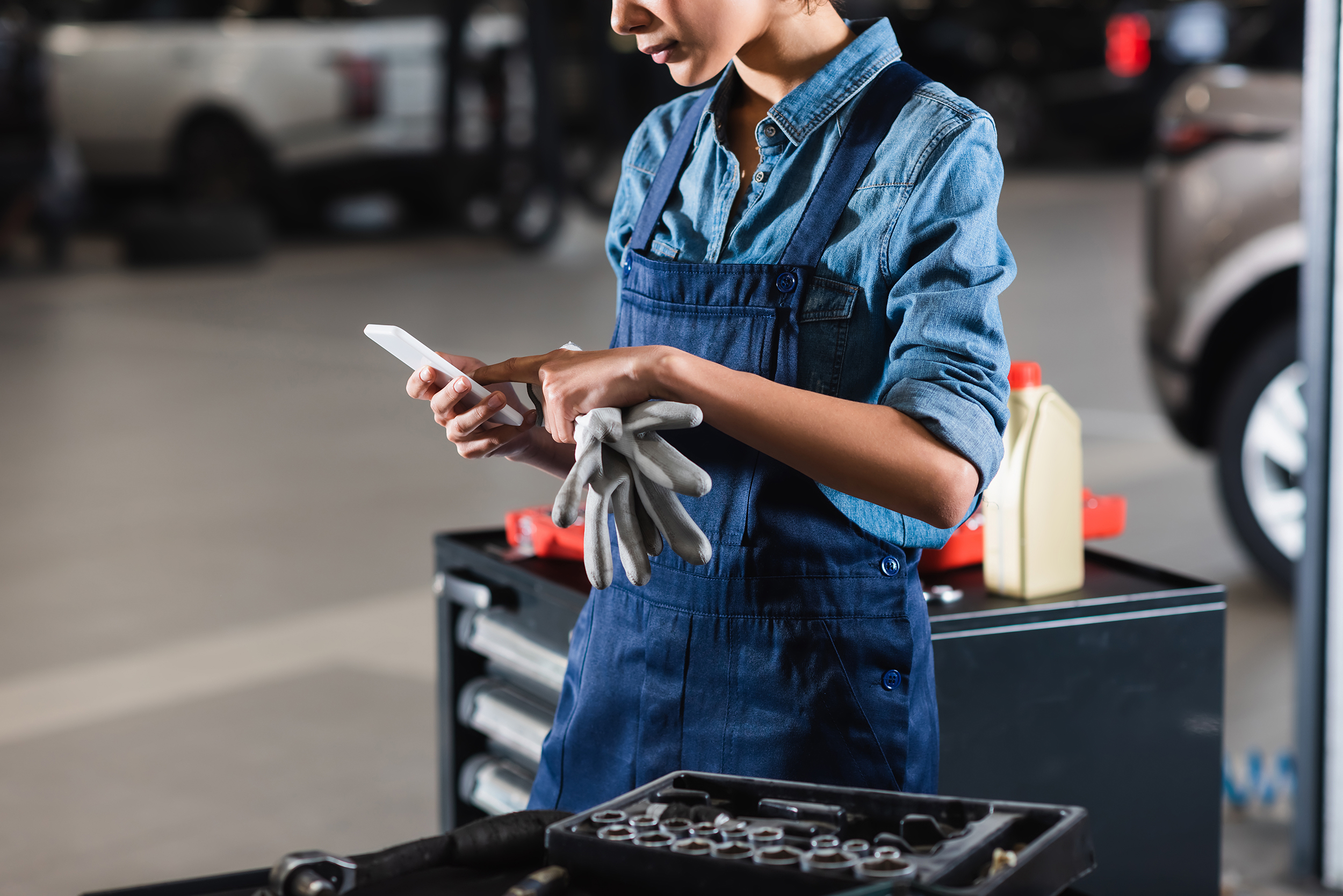 Man sieht eine junge Frau in blauem Overall, die in einer Auto-Werkstatt steht. Sie hält Arbeitshandschuhe in der Hand und tippt auf ihrem Smartphone. Im Hintergrund sind ein Werkzeugwagen und zwei graue Fahrzeuge zu erkennen.