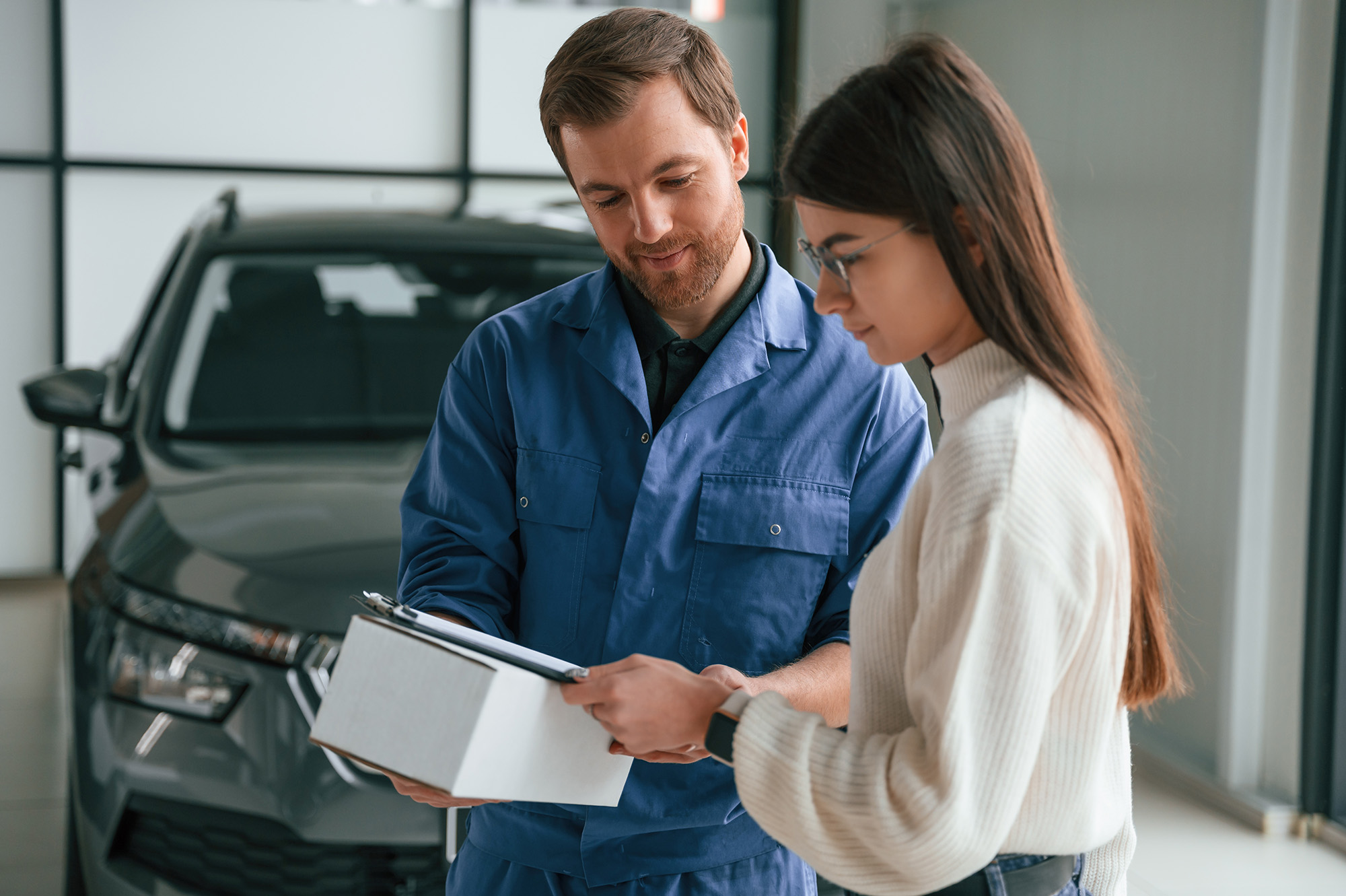 Ein junger Werkstatt-Mitarbeiter in blauem Overall berät eine Kundin der Generation Z. Sie sind einander leicht zugewandt und sehen auf ein Klemmbrett. Im Hintergrund ist ein graues Auto zu erkennen.